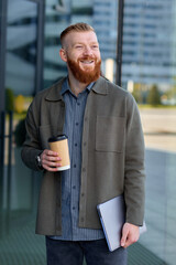 A European man with a red beard dressed in a shirt and jacket walks around the city and drinks coffee during his lunch break