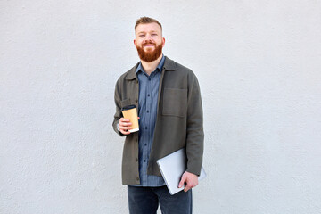 A 35 year old man with a red beard walks around the city and drinks coffee during his lunch break in the office