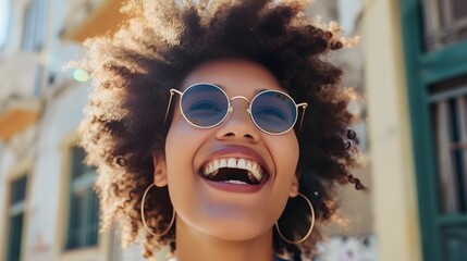 Cheerful young woman with afro hairstyle wearing sunglasses and hoop earrings, laughing happily and looking at the camera.