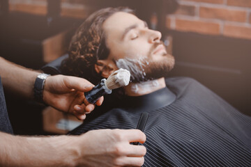 Closeup brush and razor in hands of barber, background client is waiting for shaving his beard and...