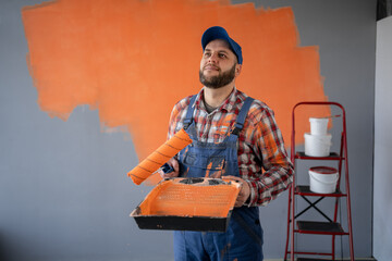 Painter man holding a paint roller and plastic tray on orange wall background thinking an idea...