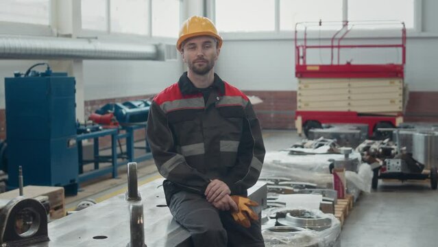 Medium portrait of young Caucasian male employee in workwear and hard hat sitting on workbench inside industrial factory and posing for camera
