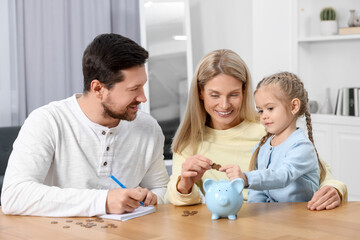 Fototapeta premium Planning budget together. Little girl with her family putting coins into piggybank at table indoors