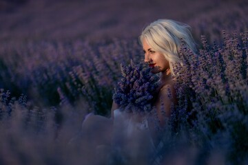 Blonde woman poses in lavender field at sunset. Happy woman in white dress holds lavender bouquet....