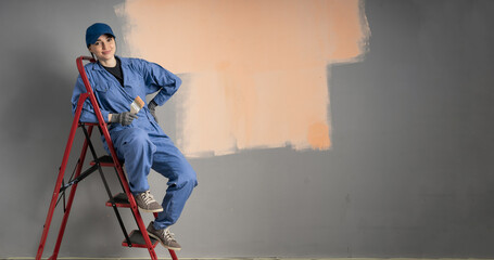 Beautiful young woman sitting on ladder doing wall painting and looking at camera. Space for text
