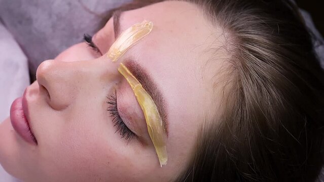 The waxer spreads the wax over the lower contour of the eyebrow to remove excess hairs. Permanent makeup procedure, performing PMU of eyebrows