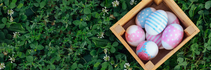 Banner. Multicolored eggs lie in a wooden box on the grass in the garden on a sunny spring day....