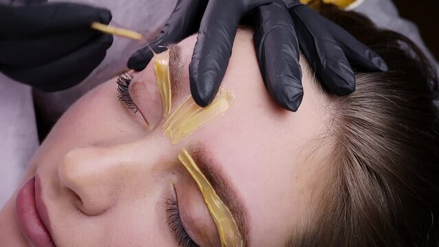 Applying yellow-colored wax to the lower contour of the eyebrow to remove hairs. Permanent makeup procedure, performing PMU of eyebrows