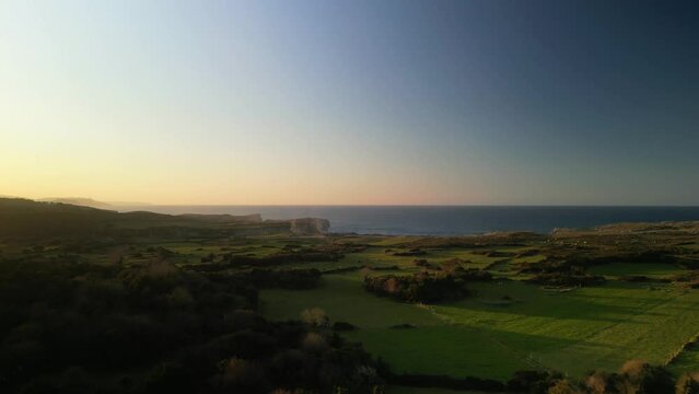 Vista a&eacute;rea de un atardecer de unas praderas vi&eacute;ndose el horizonte naranja al fon con el mar