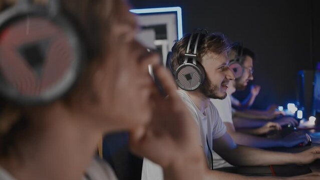 Bristly adult gamer in headset emotionally smiles and plays video game on PC at computer club, cyber sport tournament. E-sport championship. Five players team. Multiplayer online shooter, strategy RTS