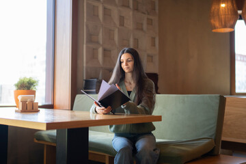 Young Woman Seated on a Green Sofa Choosing a Meal From a Menu in cafe