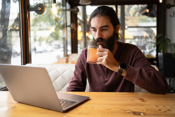 A young man with a long beard and hair working remotely as a freelancer uses a laptop, drinks tea while enjoying a break from work. Freelancing