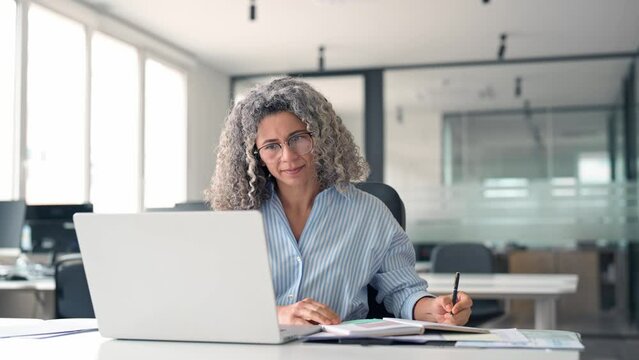 Busy mature business woman working in office using laptop writing. Smiling middle aged professional executive looking at computer, elearning, watching webinar having hybrid meeting sitting at desk.