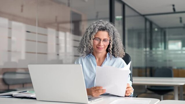 Busy middle aged business woman working in office with laptop reading document. Mature professional hr manager, notary holding paper finance report sitting at desk in office. Consultancy.
