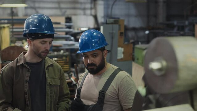 Medium shot of two multiracial male employees wearing blue hard hats standing by working lathe machine at production plant and talking