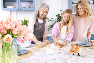 Three generations of women gather in cozy kitchen, making and roll out dough together with smiles and laughter. Grandmother teaches her daughter and granddaughter her dough recipe