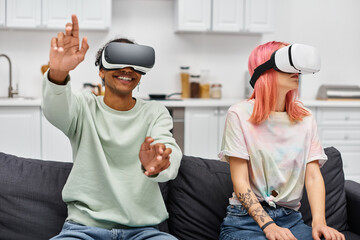 joyous beautiful multiracial couple in homewear sitting on sofa with virtual reality headsets