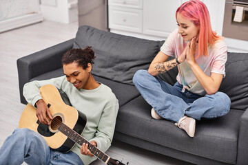 jolly woman with pink hair looking at her cheerful african american boyfriend playing guitar