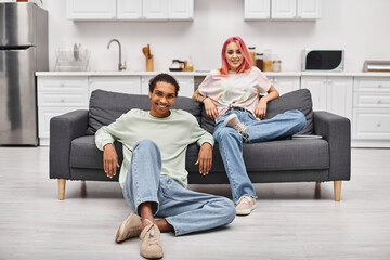 joyous attractive multiracial couple in cozy homewear posing in living room and smiling at camera