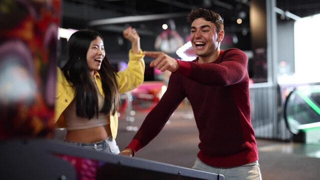 Multiracial couple of friends playing pinball in arcade game room celebrating victory in the game with jumping and hugging