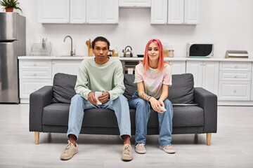cheerful multiracial couple in casual attires sitting on sofa and looking at camera at home