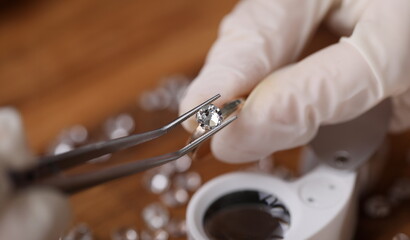 Close-up view of persons hands in protective gloves holding special equipment and examining quality of diamond. Magnifying loupe on table. Luxury jewelry concept
