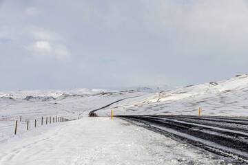 Obraz premium Panoramic low angle winter landscape in Iceland with white clouds over snowy and icy hilly landscape partly cleaned road winding through the landscape