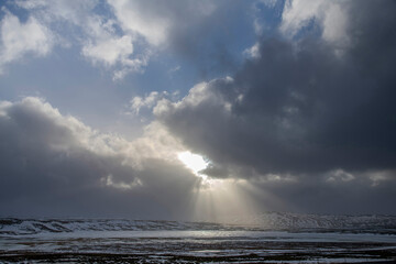Dramatic winter landscape in Iceland with white and black clouds over snowy and icy hilly landscape with bright sun between clouds and sunrays on shining on white snow landscape