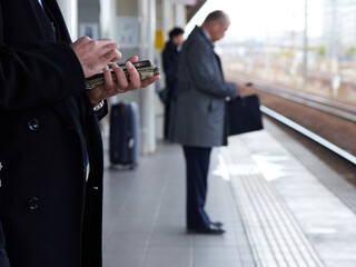 冬の駅でスマホを見ながら電車待ちの中高年サラリーマンの姿 - obrazy, fototapety, plakaty