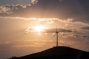 Aerogenerador en el horizonte sobre una colina al atardecer. el sol al fondo en un cielo con nubes...