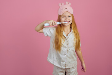 Obraz na płótnie Canvas Smiling redhead teen girl holding toothbrush and wearing sleeping mask on pink background.