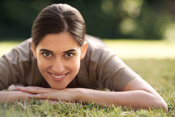 Portrait, Indian woman and lying on grass with smile at park for joy, refresh and relax with me time. Female person, bokeh and happy for leisure with break for rest, peace and quiet with wellbeing