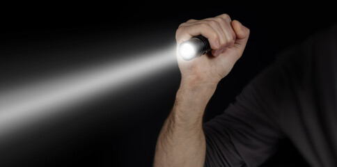 Male hand holding a led flashlight with a wide white beam on a black background, leaving the right...