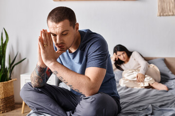 young depressed tattooed man sitting on bed near offended asian wife, family divorce concept