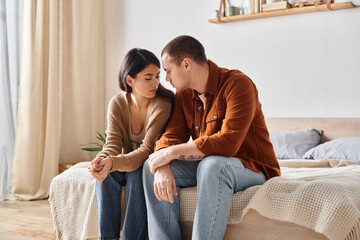 young and sad interracial couple sitting and talking on bed at home, relationship difficulties