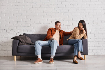 young man calming upset asian wife sitting on couch in living room, relationship difficulties