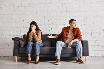 discouraged man pointing at offended asian wife on couch in living room, relationship difficulties