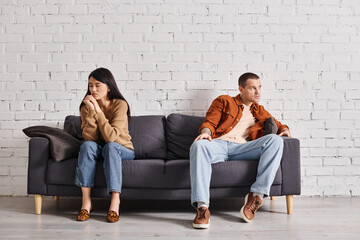 depressed interracial couple looking away while sitting on couch in living room, divorce concept