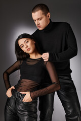 seductive asian woman in black leather pants looking at camera near handsome man on grey backdrop