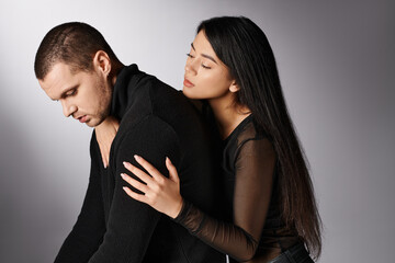 loving asian woman embracing young upset man sitting on grey backdrop, affection and fashion