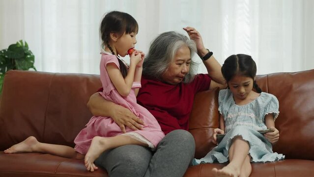 Footage handheld medium shot, Grandmother sitting on sofa and hugging little girl eating strawberry and grandma have fun talking with granddaughter who playing with tablet in living room