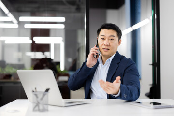 Fototapeta premium Adult entrepreneur having phone conversation while sitting bu equipped table with computer in corporate building. Calm startup leader discussing project details with perspective investor on distance.
