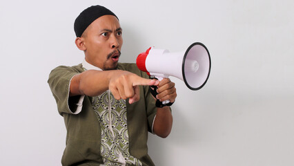 Angry Indonesian Muslim man in koko and peci shouts into a megaphone, pointing his finger at the...