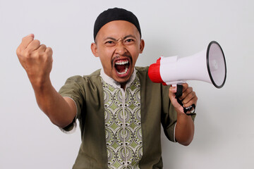 Angry Indonesian Muslim man in koko and peci shouts in protest, raising his fist in defiance while...