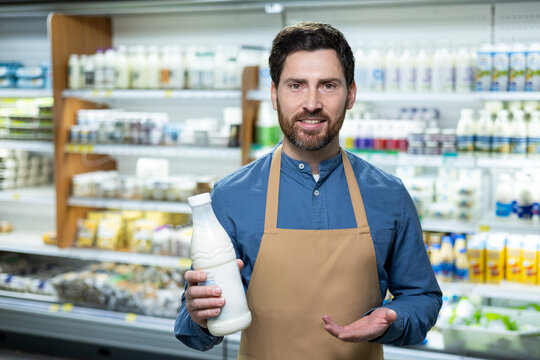 Experienced male employee in an apron stands in the supermarket's dairy section, suggesting a selection of milk and yogurts. The image captures the essence of daily grocery shopping and helpful