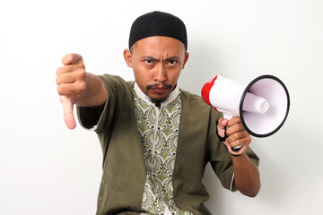 Indonesian Muslim man in koko and peci gives a thumbs-down gesture while holding a megaphone,...