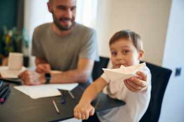 Playing with origami boat. Father and son are at home by the table