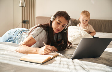 Woman in headphones with laptop, writing in notepad. Mother with her little son is at home in...