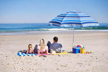 Family, relax and lying on beach with umbrella for summer holiday, vacation or outdoor weekend together in nature. Rear view of father, mother and daughter chilling on towel or sand by ocean coast