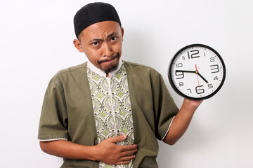 Hungry Indonesian Muslim man in koko and peci holds his stomach and looks longingly at a clock,...